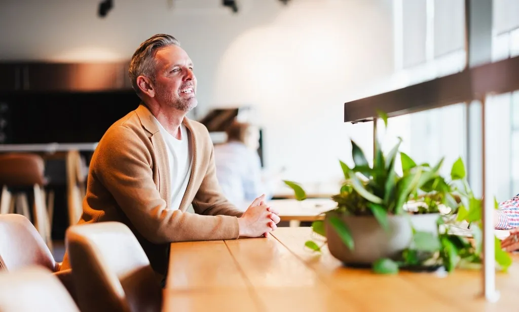 A professional man in a tan cardigan smiling thoughtfully while sitting at a wooden table in a bright office.